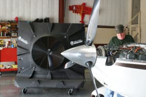 A person works on a small aircraft engine in a hangar. Nearby, a large industrial fan is positioned. The hangar wall is visible in the background, with various tools and equipment on shelves.