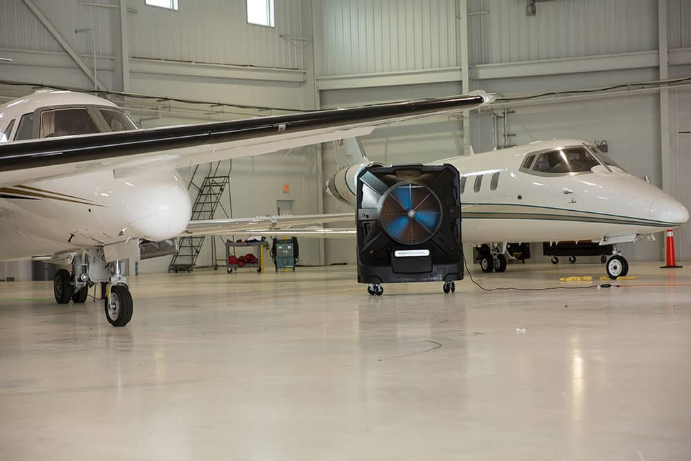 Two white private jets are parked inside an aircraft hangar. A large industrial fan is positioned between them on the hangar floor. The hangar is spacious with a clean, industrial appearance, and the planes have sleek designs.