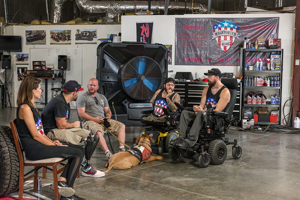 Five military veterans from Warfighter Made, two in wheelchairs, are sitting in a workshop with a Portacool in the background.