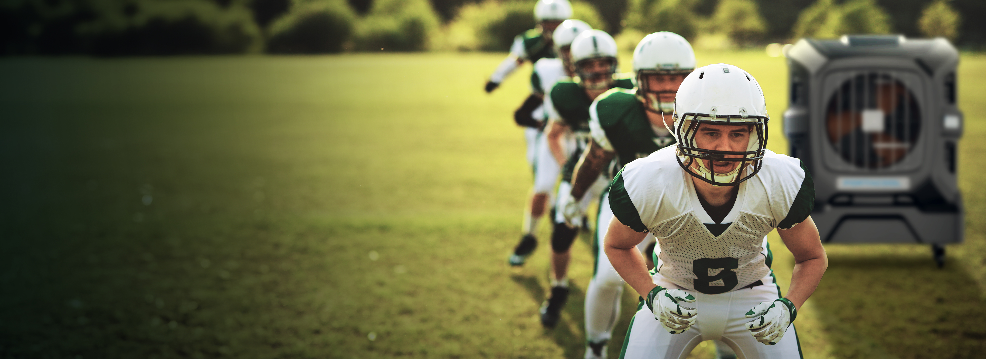 Football players in white and green uniforms prepare on the field. Player 6 leads, helmet on. A large metallic crate sits on the grass in the background.