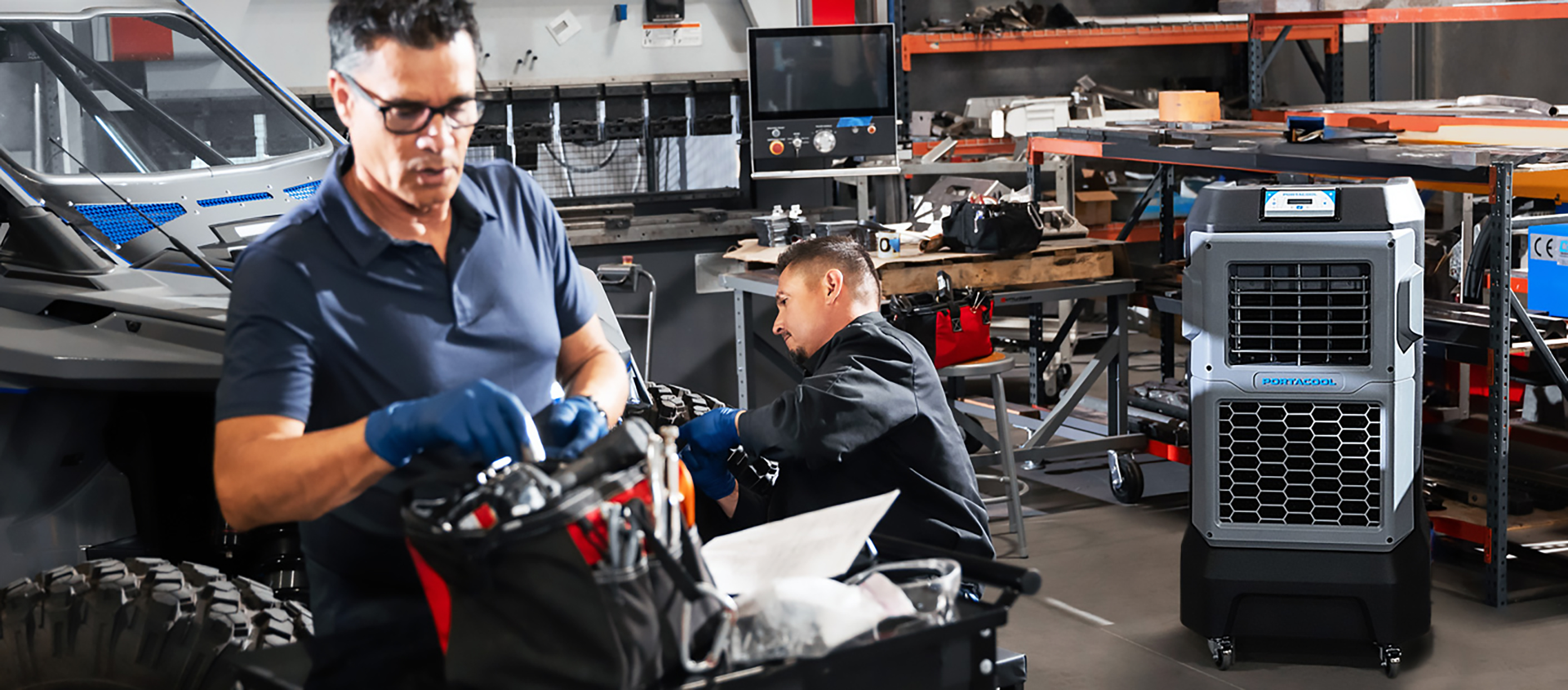 Two men work in a garage. One organizes a tool bag; another kneels by a workbench surrounded by scattered tools. A Portacool air cooler runs nearby.