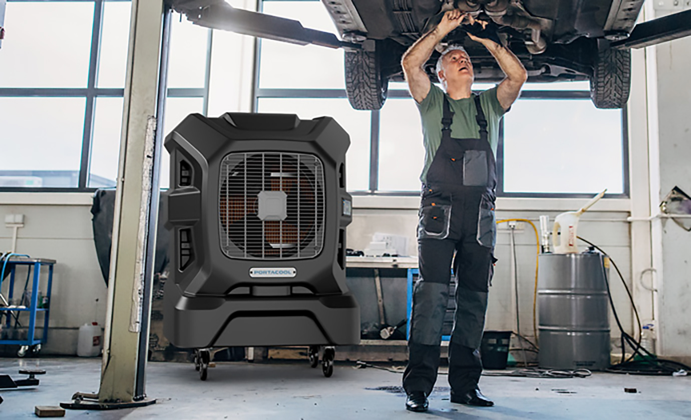 A mechanic in overalls works under a lifted car in a tidy, well-lit garage. Tools and equipment are neatly organized around the workspace.