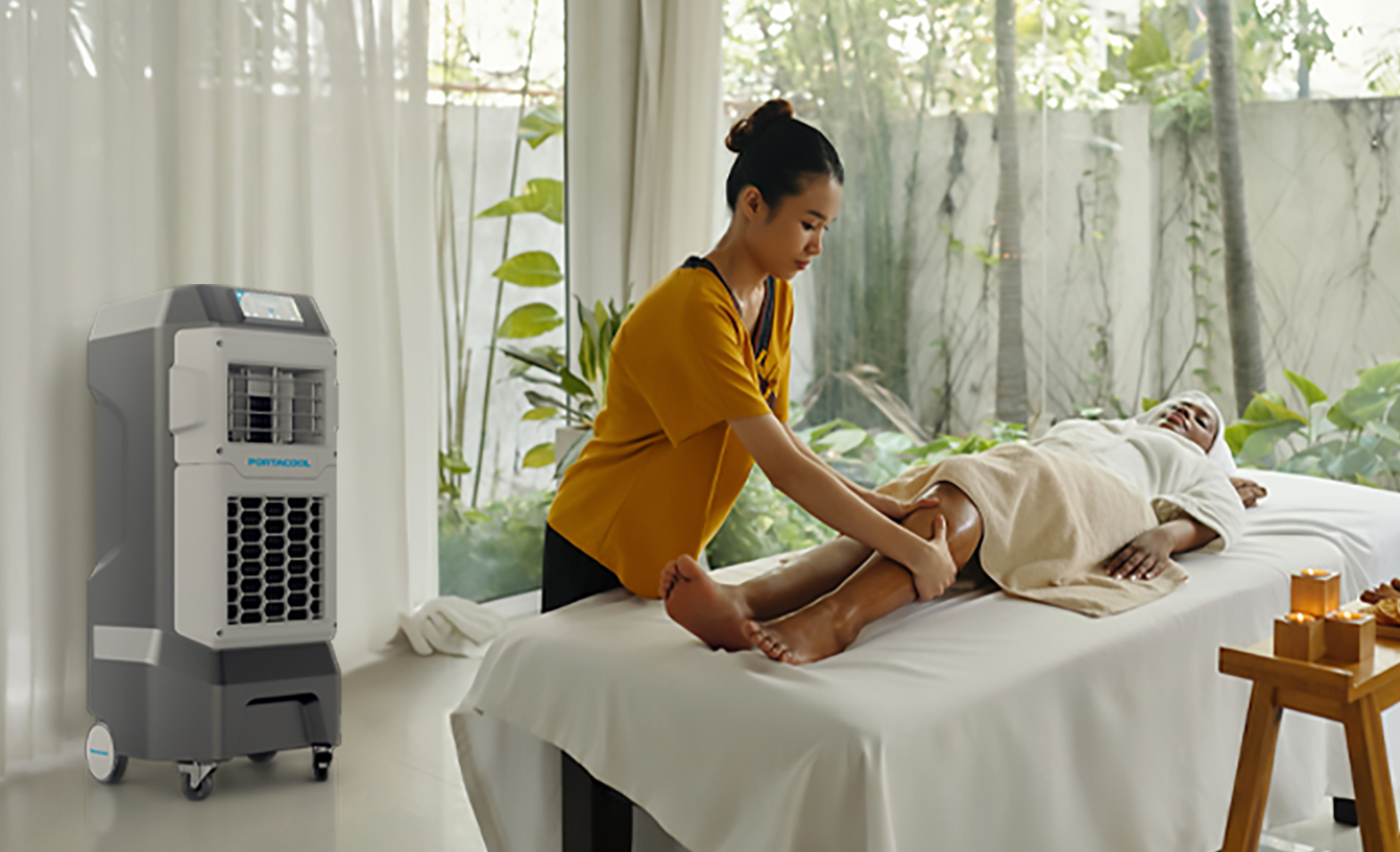 A person receives a leg massage in a serene spa with large windows, sheer curtains, greenery views, and candle-lit ambiance feeling both outdoors and cool enough for comfort.