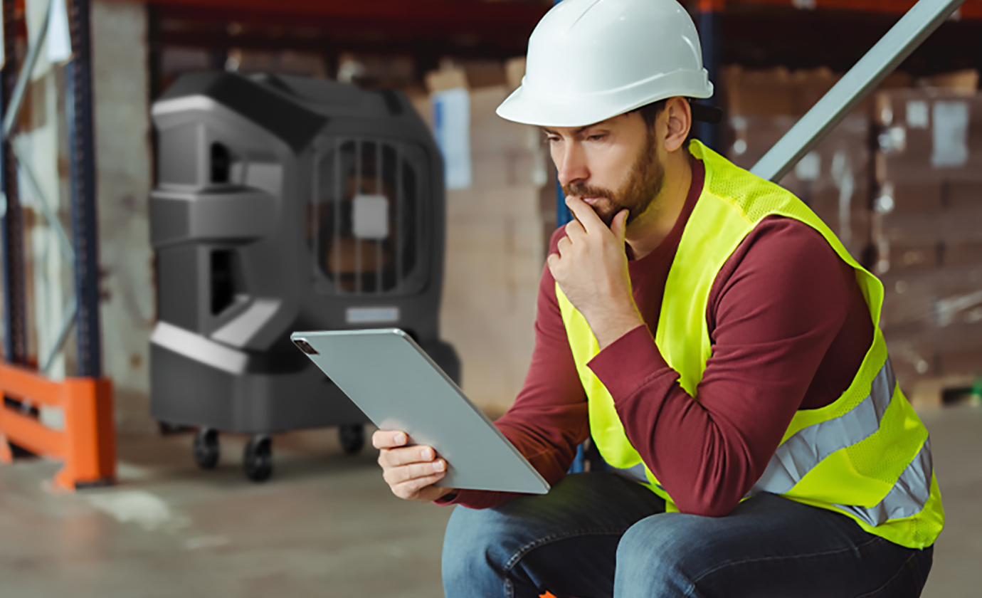 A construction worker in a vest and hard hat sits in a warehouse, focused on a tablet. Stacks of boxes and equipment are visible behind him.