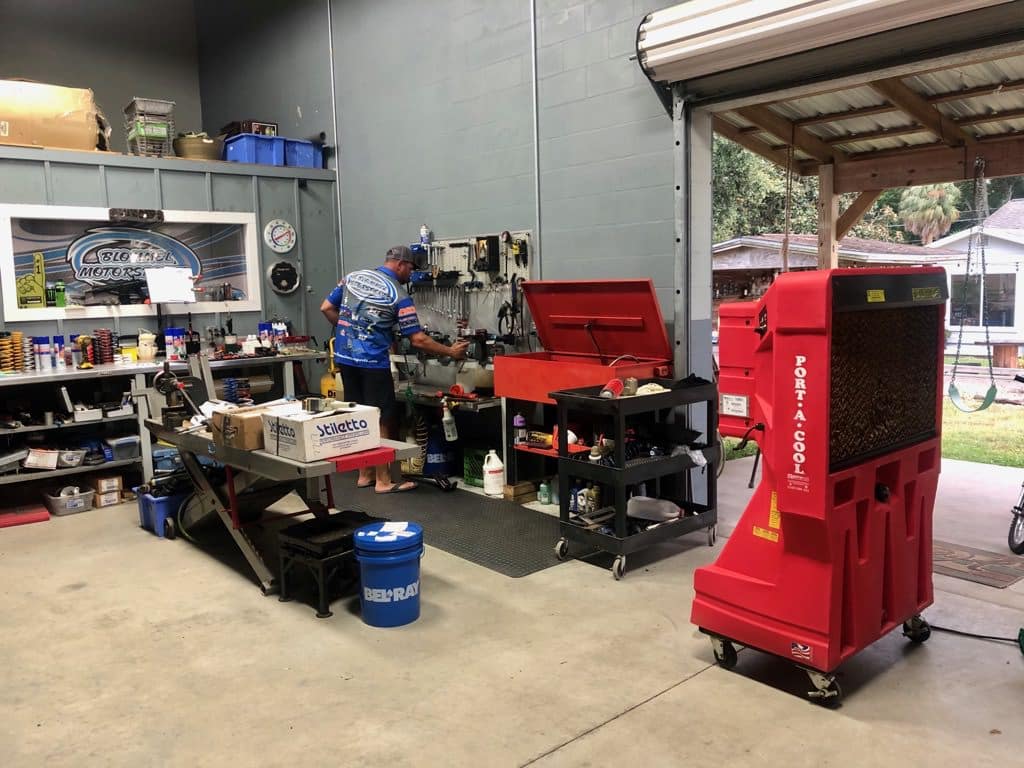 A mechanic at Blommel Motorsports works near tools and a red tool chest. A red portable cooler is nearby, with shelves and an open garage door.